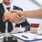 Shaking hands while wearing suits, with documents, calculator and a miniature house on the table