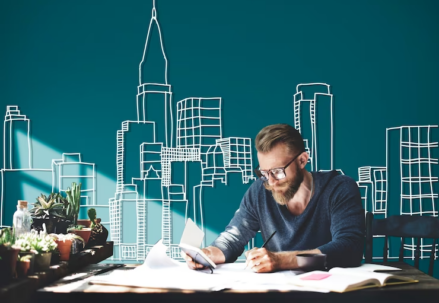 Man wearing glasses, holding a notebook in one hand and writing with the other hand, with a backdrop of buildings