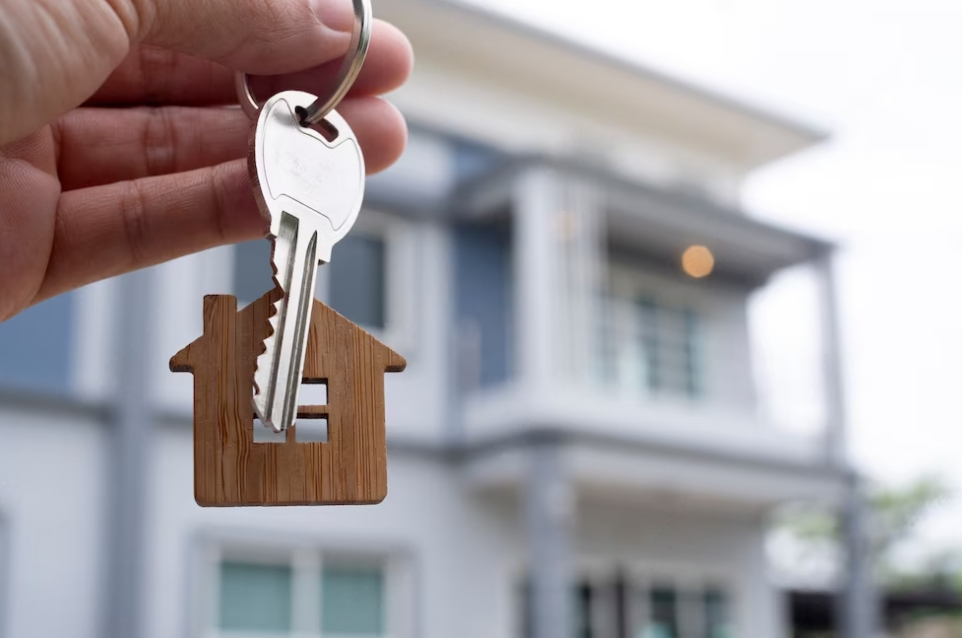 hand holds a keys with wooden house  in front of blurred house