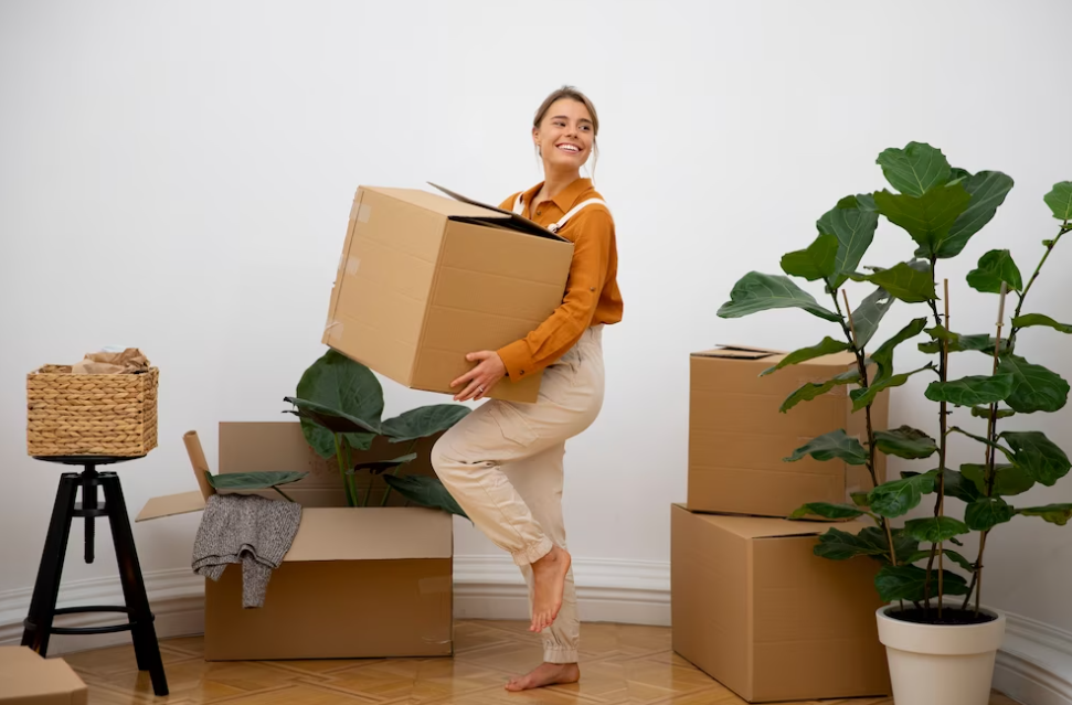 woman smiling and holding the boxes in the room with other boxes and flowers