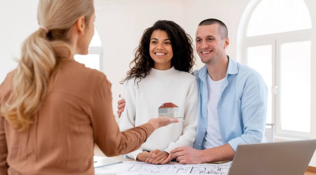 woman in brown shirt holds a small house in her hand, a couple watching on her and smiling